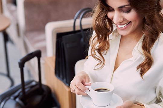 Frau mit Tasse Kaffee in der Hand, warten mit einem schwarzen Koffer neben sich 